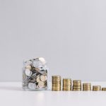 glass-jar-full-of-money-in-front-of-decreasing-stacked-coins-against-white-background